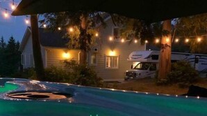 A swimming pool with a hot tub under a covered area, surrounded by trees and a house with string lights. - Viceroy home at Soames Hill (Gibsons)