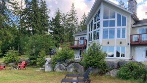 A two-story house with a large deck and a stone fireplace in the yard. - Viceroy home at Soames Hill (Gibsons)