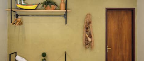 A bathroom with a wooden shelf holding a yellow boat model, a potted plant, and a hanging nest decoration. There is a toilet, a towel, and a small plant on a lower shelf.