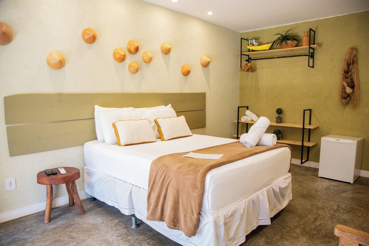 Une chambre d’hôtel comprenant un lit, des tables de chevet, un petit tabouret et une étagère fixée au mur, agrémentée d’objets décoratifs.