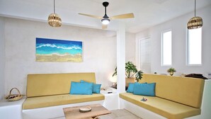 A modern living room with two yellow sofas, a wooden coffee table, a ceiling fan, and a beach-themed painting on the wall. - 2BR - Fast Internet, Near Beach, Pool, King Beds, ACs (Puerto Escondido)