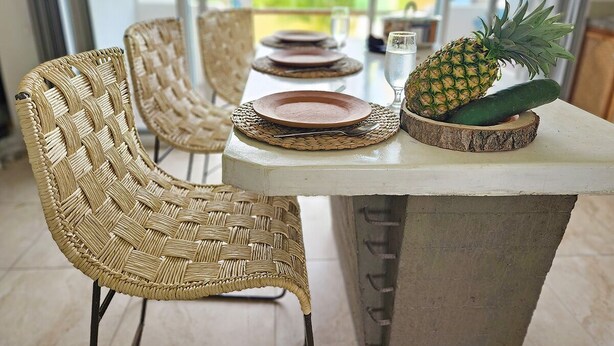 A dining area with wicker chairs, a table with plates and a pineapple, and a view through large windows. - 2BR - Fast Internet, Near Beach, Pool, King Beds, ACs (Puerto Escondido)