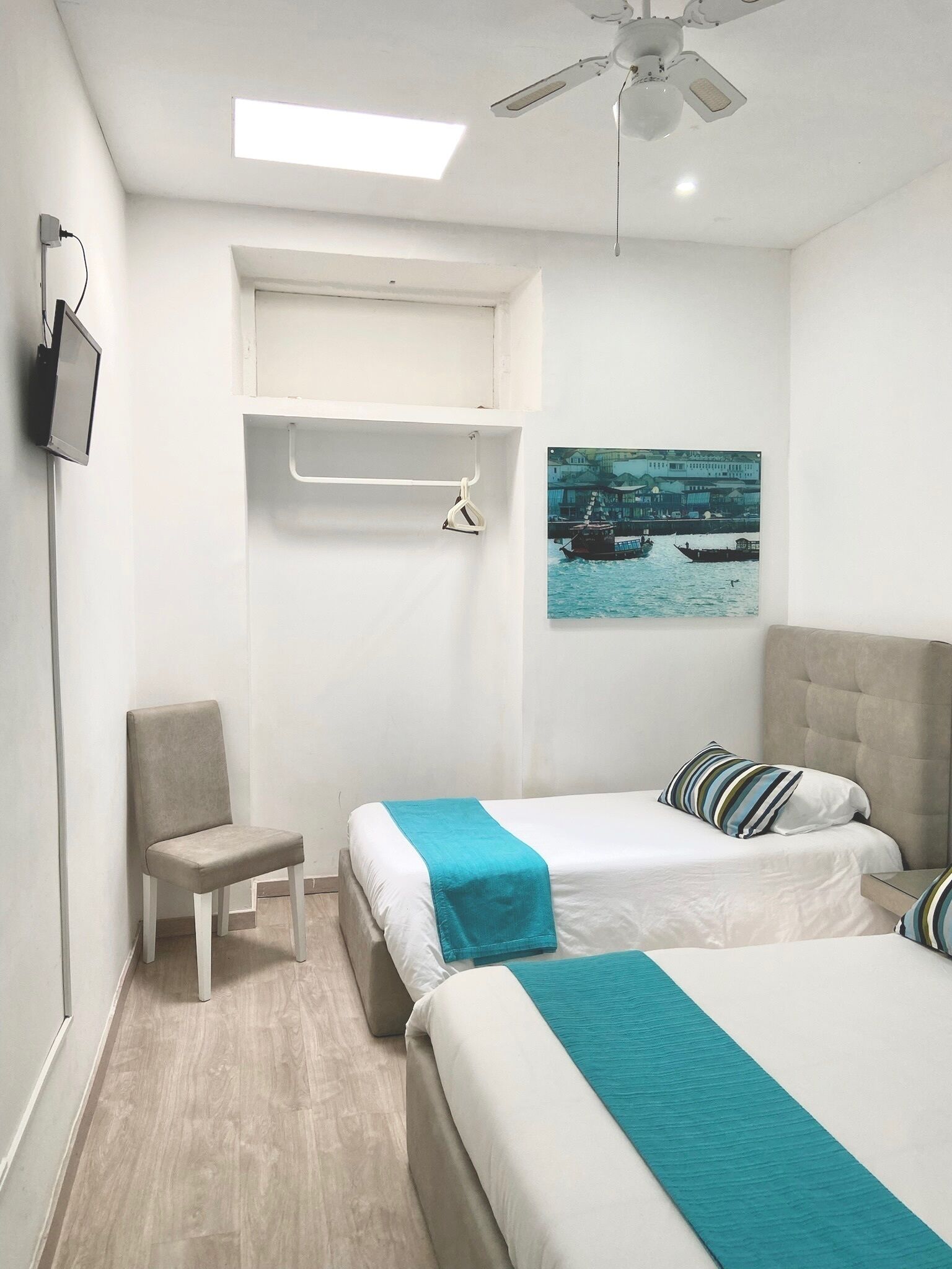A hotel room with two beds, a chair, a TV, a ceiling fan, and a painting of a boat on the wall.