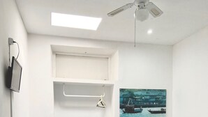 Ein Hotelzimmer mit zwei Betten, einem Sessel, einem Fernseher, einem Deckenventilator und einem Gemälde eines Bootes an der Wand.
