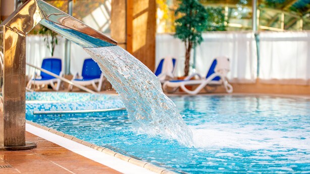 An indoor swimming pool with clear water, a metal water feature, and lounge chairs in the background. - Alga Baltic Resort (Swinoujscie)