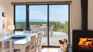 Un área de comedor con mesa y sillas, una chimenea y un amplio ventanal corredizo que da a una terraza exterior con vistas a las montañas.