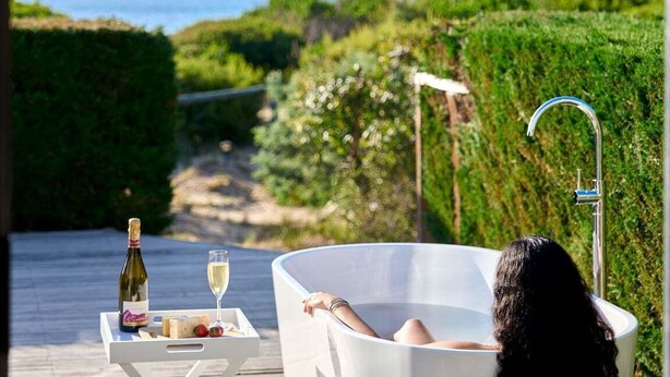 Una persona relajándose en una bañera con una botella de vino y una copa sobre una mesa, con vistas a un jardín y al mar.