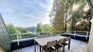 En terrasse med et bord, stole, gelænder og udsigt til et hus og træer.