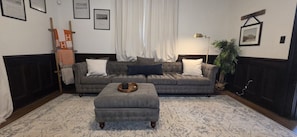 A living room with a grey sofa, a matching ottoman, a patterned rug, and a lamp.