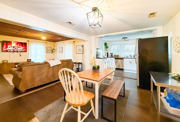 A modern living room with a sofa, a dining table with chairs, a kitchen area, and a large TV.