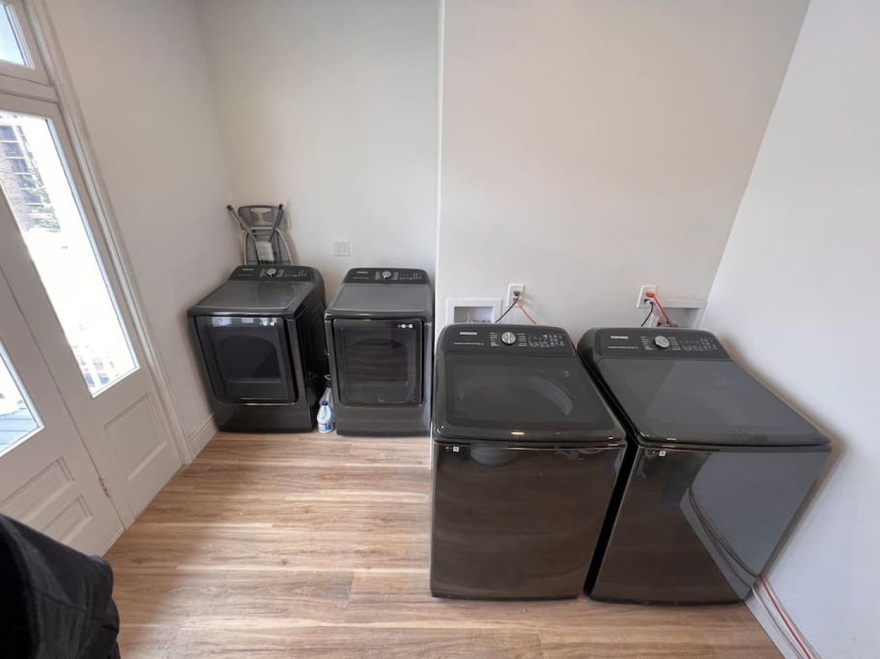 A laundry room with four washers and dryers, wooden flooring, and a window providing natural light.