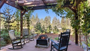 En träveranda med en eldstad och fyra gungstolar, omgiven av träd och en pergola med ljusslingor.