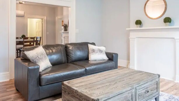 A modern living room with a dark leather sofa, a wooden coffee table, and a white fireplace mantel with two small potted plants. - Modern Modest Design  Newburgh, Beacon, Westpoint  2nd. FL Apt. (Newburgh)
