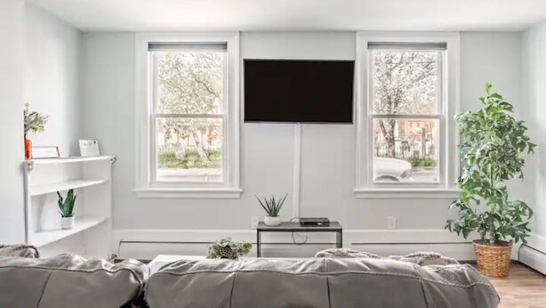 Une pièce dotée d’une grande fenêtre, d’un téléviseur à écran plat fixé au mur, d’une petite table avec une plante et d’un lit avec une literie grise.