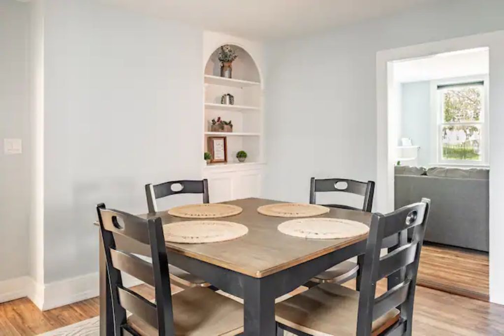 A dining area with a wooden table and four chairs, a white shelving unit with decorative items, and a view of a green outdoor area through a window.