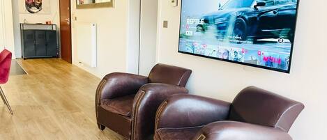 A living room with a flat-screen TV mounted on the wall, leather armchairs, a wooden floor, a radiator, a cabinet, and a framed artwork on the wall.