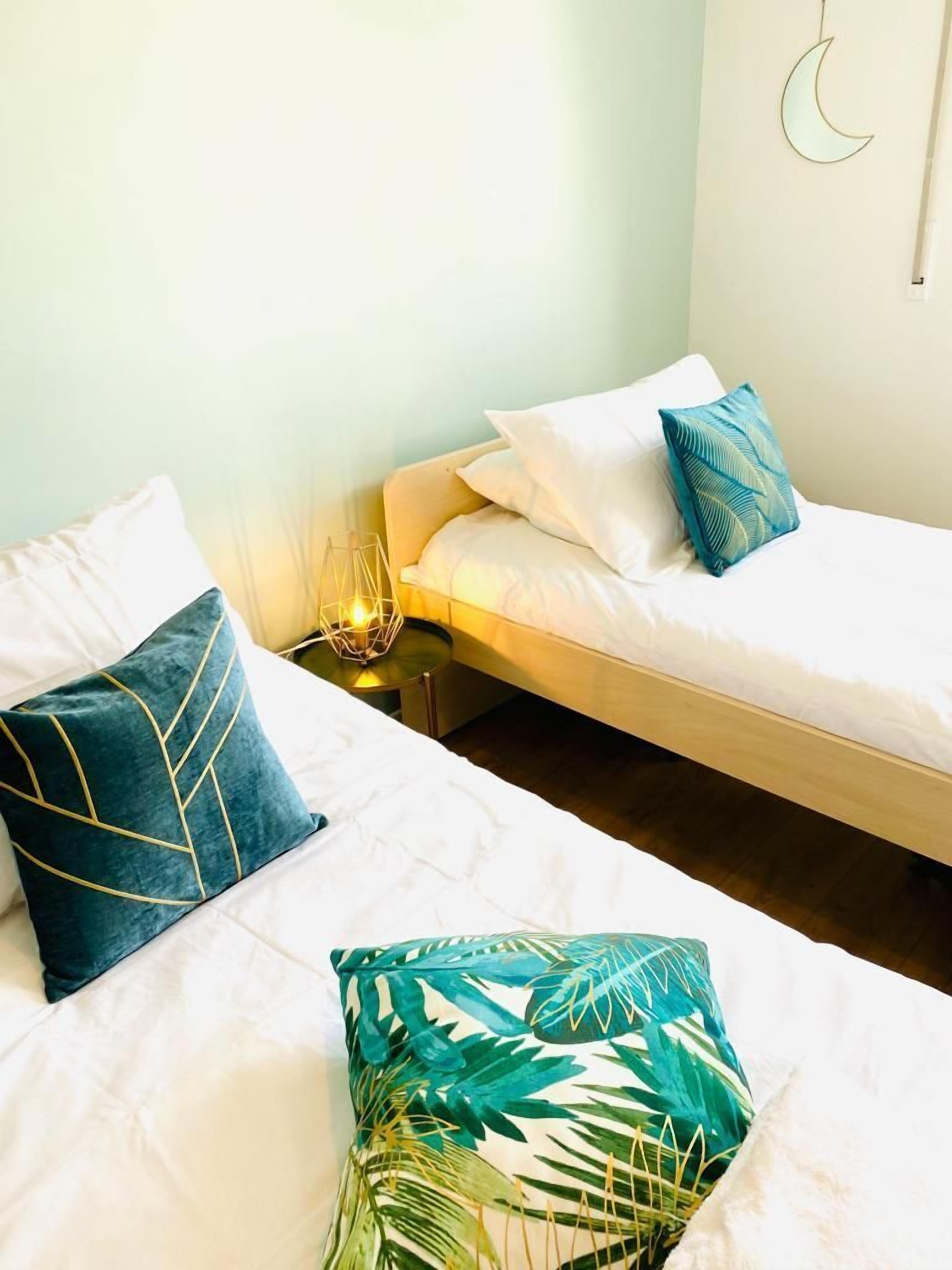 Quarto com duas camas, cada uma com roupa de cama branca e almofadas decorativas. Há uma mesinha de cabeceira com abajur entre as camas.