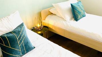 Quarto com duas camas, cada uma com roupa de cama branca e almofadas decorativas. Há uma mesinha de cabeceira com abajur entre as camas.
