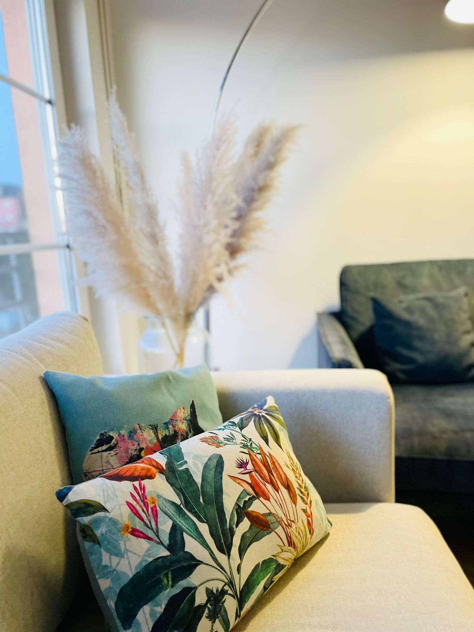 Una sala de estar con un sofá, un cojín decorativo con estampado tropical y una alta jarra con plumas de pampa.