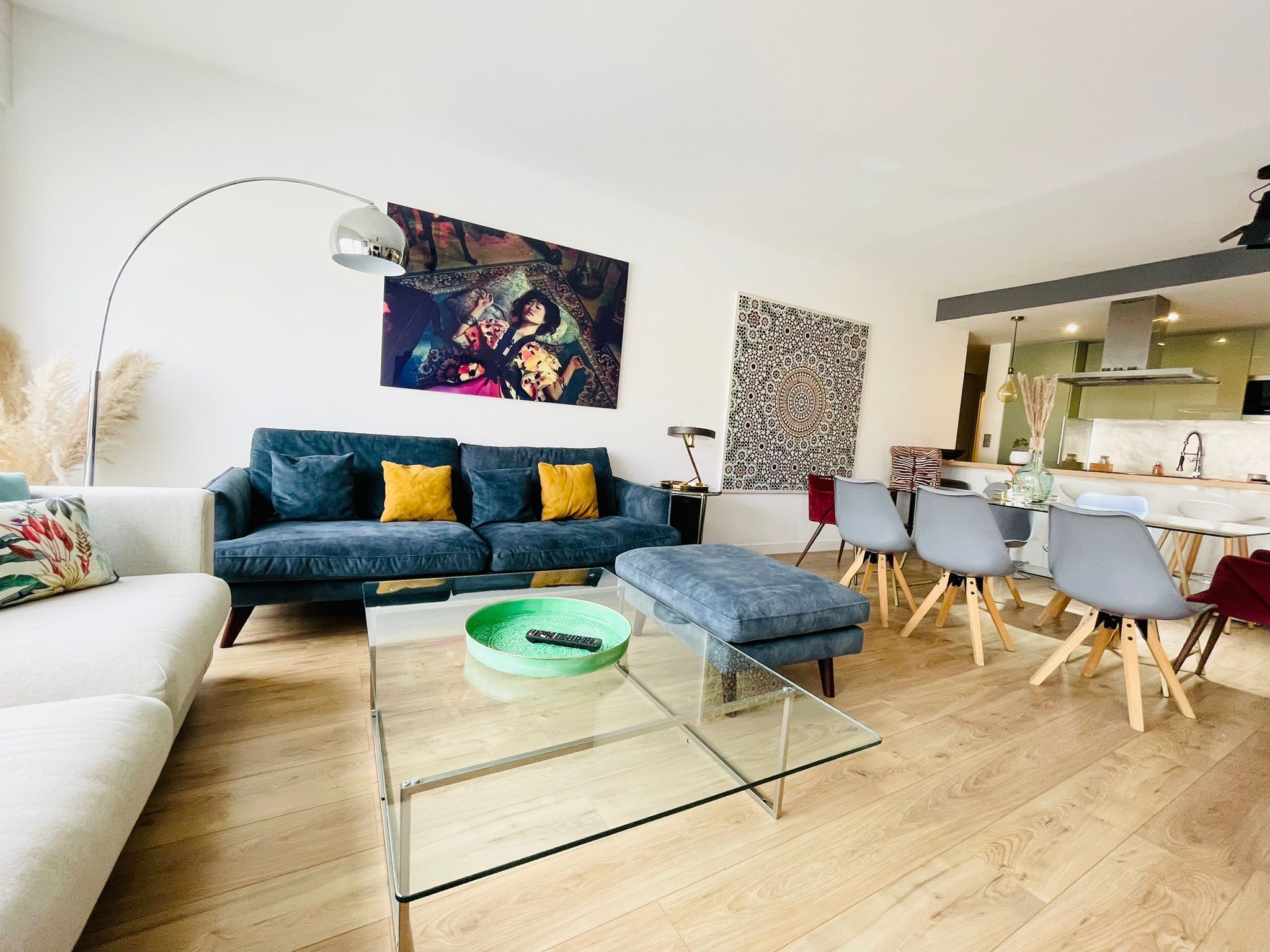 A modern living room with a sofa, ottoman, and glass coffee table. There is a dining area with chairs and a kitchen in the background.