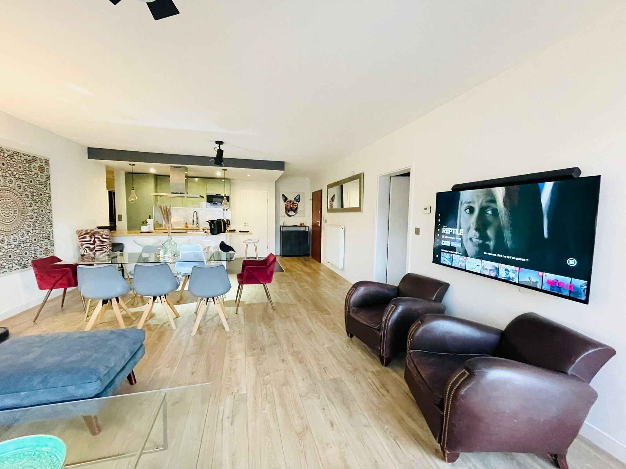 A modern living room with a dining area, a kitchen in the background, and a flat-screen TV mounted on the wall.