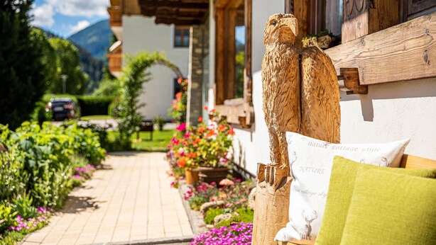 A rustic wooden bench with a cushion, a wooden sculpture, and a garden path leading to a building with a wooden facade and a window with a curtain.