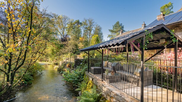 A riverside garden with a stone wall, a wooden deck, and a pergola with vines. - The Waterfall Retreat (Penrith)
