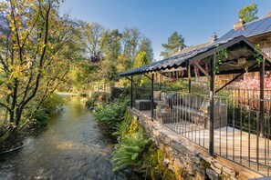 A riverside garden with a stone wall, a wooden deck, and a pergola with vines. - The Waterfall Retreat (Penrith)