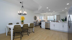 A modern kitchen with white cabinetry, a dining area with wicker chairs, a chandelier, and a view through large windows.
