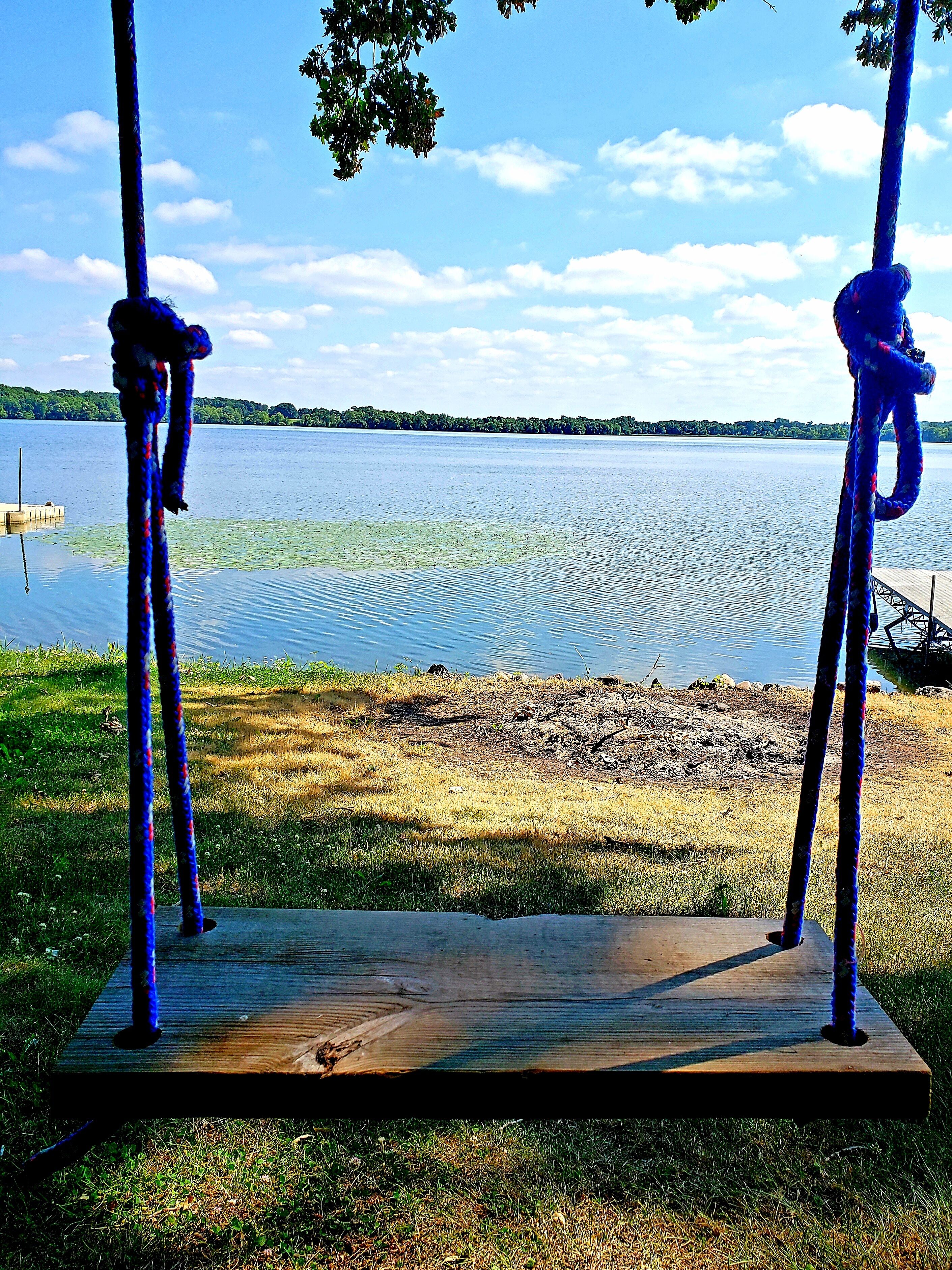 A wooden swing set with ropes, located near a lake with a dock in the background.