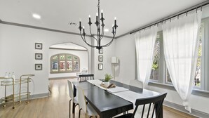 Een eetruimte met een zwarte tafel, witte stoelen, een kroonluchter en uitzicht op het landschap door grote ramen met witte gordijnen.