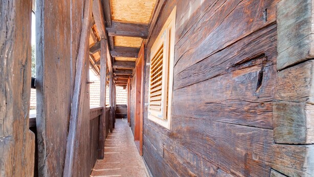 Un étroit couloir en bois, avec des murs en bois à lattes et un plancher en bois, qui mène à un espace doté de fenêtres.