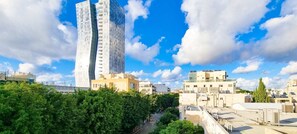 A modern urban landscape with a prominent curved skyscraper, surrounded by smaller buildings and lush green trees. - Dizengoff Muguet (Tel Aviv)