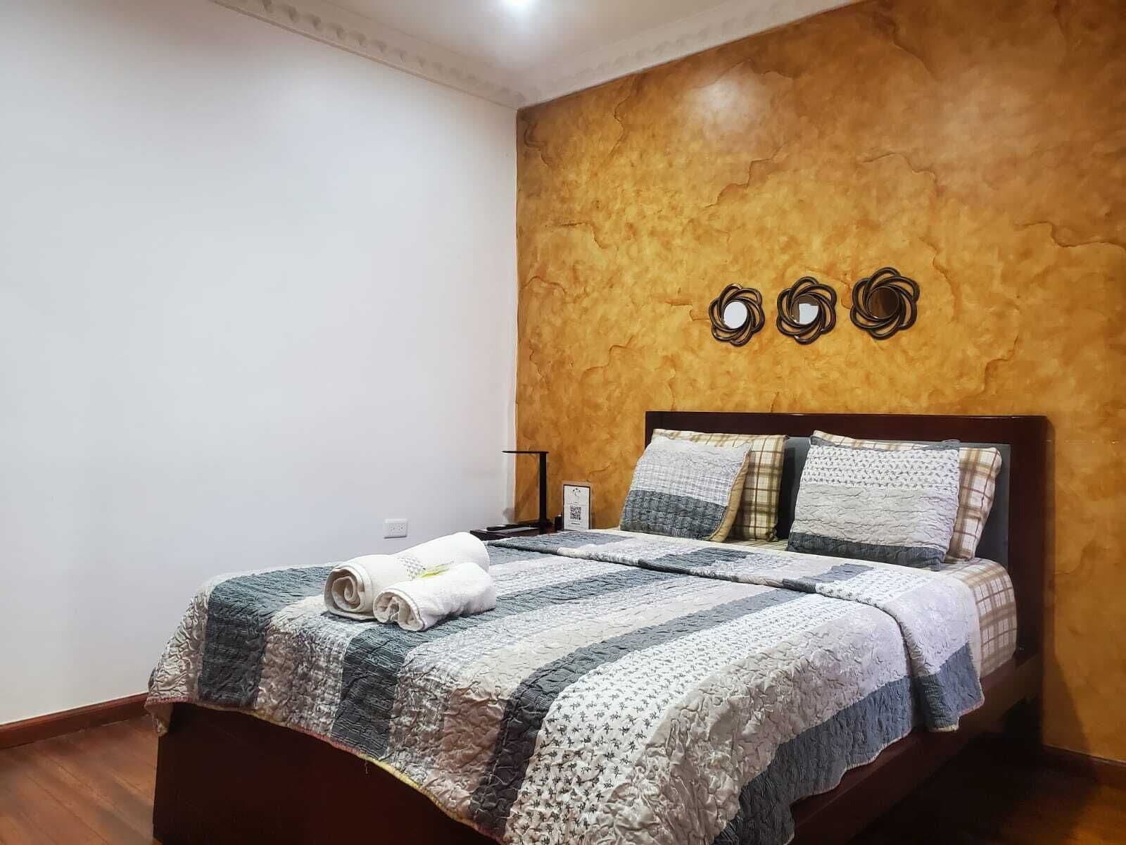 A neatly made bed with a quilt and rolled towels, a bedside table with a lamp, and a wall with three circular mirrors.