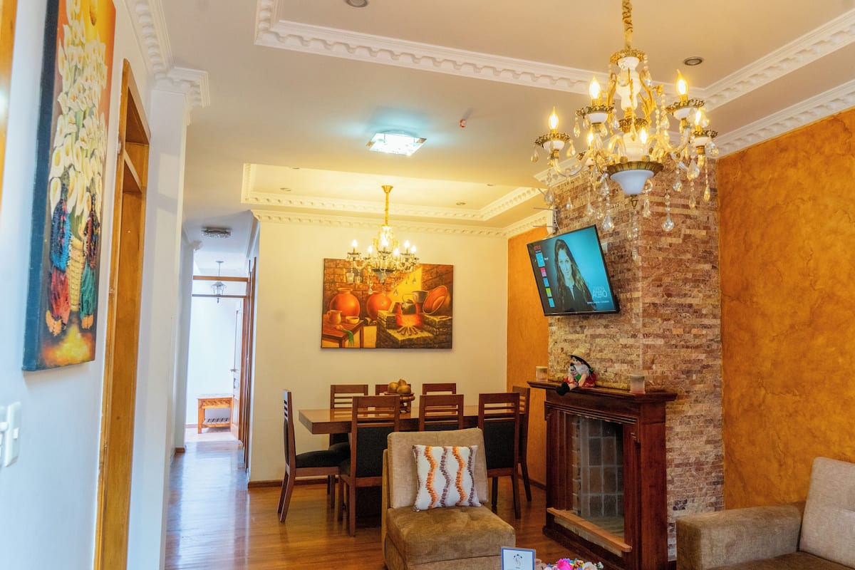 A living room with a fireplace, a dining area with chairs, a sofa, and a TV mounted on a brick wall.