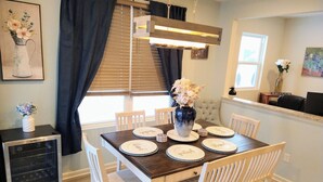 Comedor con una mesa de madera dispuesta para cuatro, sillas blancas, un florero con flores y un cuadro en la pared.