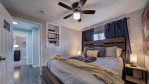 A bedroom with a bed, a ceiling fan, a nightstand with a lamp, a shelf with decorative items, and a door leading to another room.