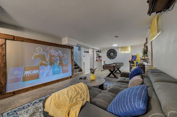Ein Wohnzimmer mit einem großen Bildschirm, der Videospieltiteln zeigt, einem Billardtisch und einer Couch mit Kissen.