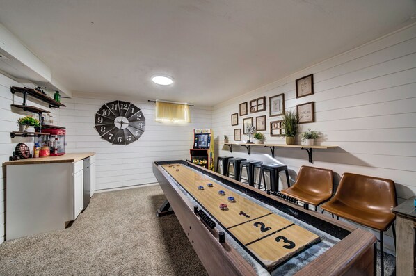 Une pièce chaleureuse avec une table de jeu de palet, une horloge et un coin bar équipé de tabourets et d’un juke-box.