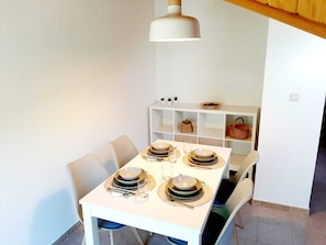 Uma sala de jantar com mesa branca posta para quatro pessoas, cadeiras, luminária pendente e um armário branco com cestos.
