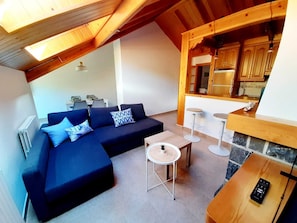 Uma sala de estar moderna com um sofá seccional azul escuro, uma mesa redonda pequena e uma área de cozinha com armários de madeira e painel de pedra.