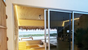 Ein modernes Gebäude mit Glastür, das zu einem überdachten Außenbereich mit Sitzbank und Blick auf den Strand führt.