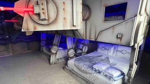 A futuristic room with a bunk bed, a small table, and a wall-mounted control panel.