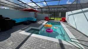A covered outdoor swimming pool with a glass ceiling, a blue sofa, red chairs, and a brick floor.