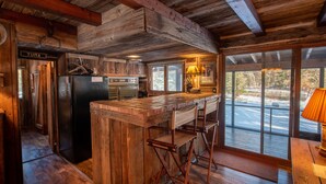A rustic kitchen with wooden beams, a refrigerator, bar stools, and a large window overlooking a snowy landscape. - Uncle Tom's 3BDR Cabin For Nature Lover (Grayling)