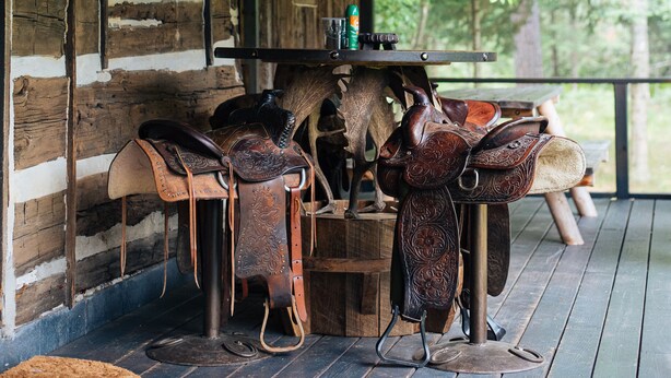 A rustic cabin interior with wooden walls, a wooden porch, and a table with leather saddles. - Uncle Tom's 3BDR Cabin For Nature Lover (Grayling)