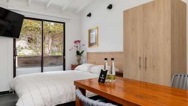 Une chambre à coucher avec un lit, une table en bois, une chaise, un téléviseur fixé au mur et une fenêtre donnant sur des arbres.