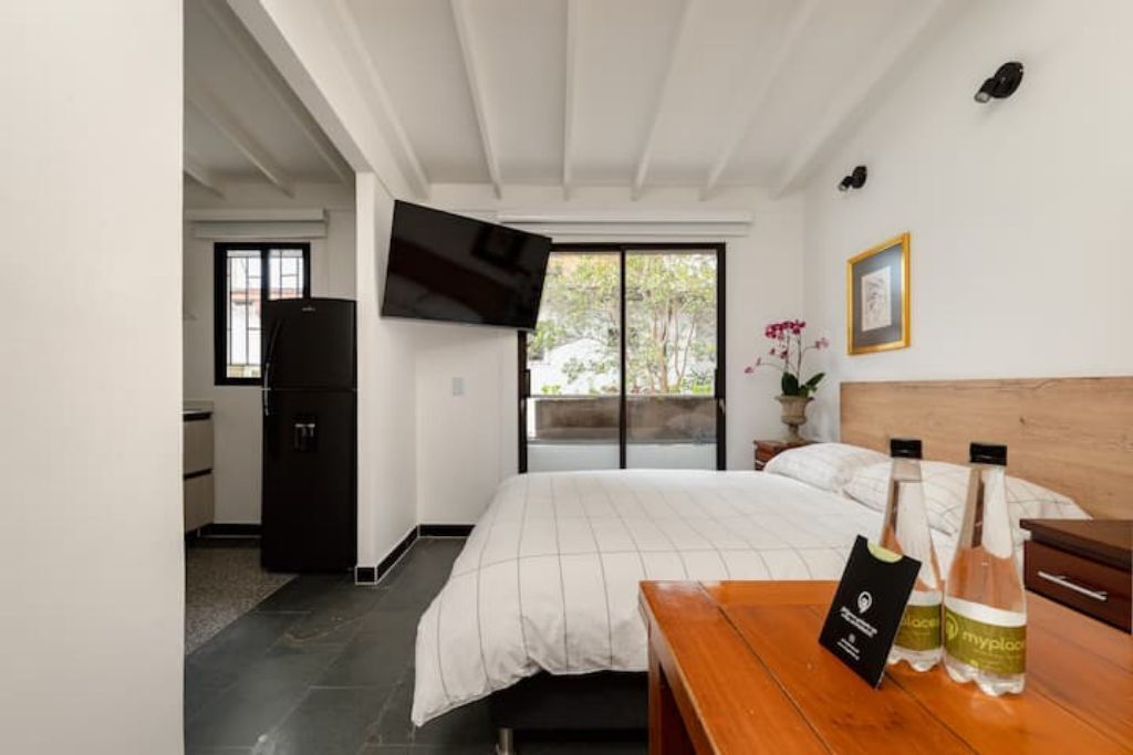 A bedroom with a bed, a wooden nightstand, a TV mounted on the wall, and a window with a view of greenery.