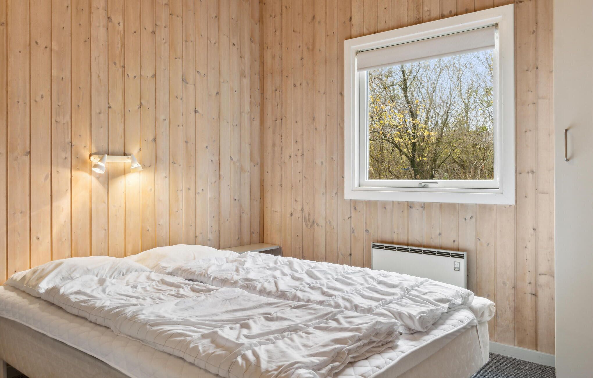 Een slaapkamer met een houten wand, een bed met wit beddengoed, een raam met uitzicht op bomen en een wandlamp.