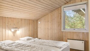 Een slaapkamer met houten wanden, een bed met witte lakens, een raam met uitzicht op bomen en een wandlamp.